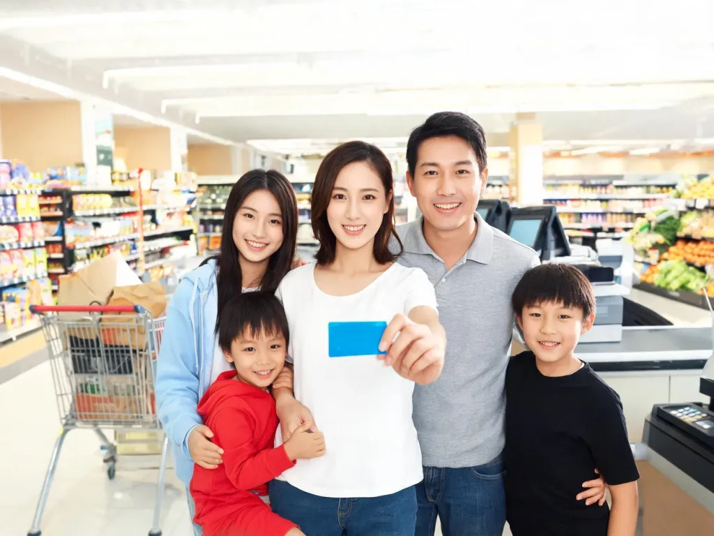 Uśmiechnięta wielopokoleniowa rodzina przy kasie w supermarkecie, rodzice pokazują niebieską kartę zniżkową.